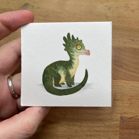 photo of a hand holding a tiny painting of a green dragon with a yellow belly and yellow eyes