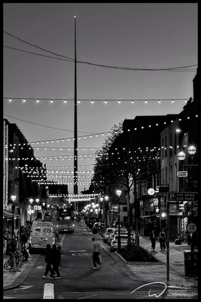 Street in Dublin North decorated with burning lights across the road as the evening falls. Shops, bikes, cars and pedestrians on the left and right. A crossroads further on stands a tall pole, The Spire, with a light on top.