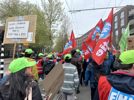 Crowd of protestors wearing green baseball caps and carrying red flags. Protest sign reads “demolish education = demolish democracy phase 1. Stop phase 1”