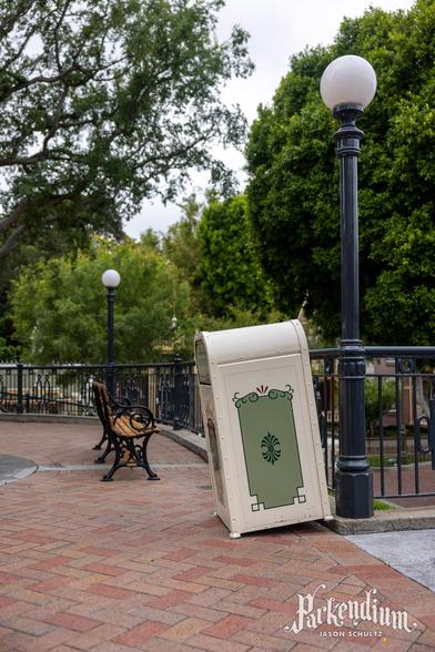 A tan trash can with decorative patterns tilted slightly next to a black iron fence.