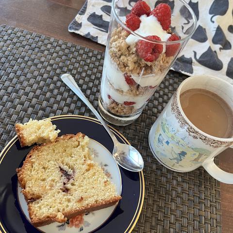A slice of cake on a decorative plate next to a long tea spoon, a parfait glass filled with layers of granola, yogurt, and raspberries, and a mug of creamed coffee on a woven placemat.
