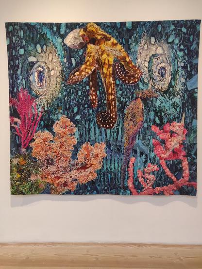 The Crepuscular Zone by Sarah Perret. A large tapestry depicting a gold and brown octopus among corals. The blue background depicts two huge, looming eyes of a predator. The whole piece is strongly textured and mosaic-like, so the predator can only be recognised from far away.