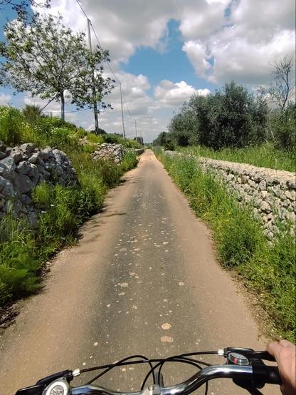 Foto mentre pedalo. Davanti: un rettilineo di una stretta strada di campagna polverosa, a sinistra della vegetazione, a destra un muretto a secco. In alto cielo azzurre e nuvole bianche
