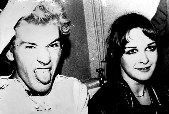 Black and white photo of Captain Sensible with a hand in his bleached hair, sticking his tongue out, next to a teenage proto-goth Jill Bryson.