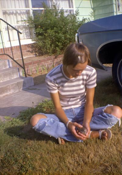 A person sits in the grass in front of a house holding a small, seemingly wild bird. They're wearing a striped t-shirt and bluejeans with large holes ripped in the knees.