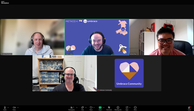 A screenshot of a Zoom meeting titled “Workplace” with five participants. Each participant is shown in their video window:

Top left: A man labeled Richard Thompson wearing a white hoodie and headphones, sitting in a softly lit room.

Top center: Owain Jones smiling, with a custom virtual background featuring the UmbraCymru logo, a map of Wales, a rocket icon, and logos for Method4 and Umbraco.

Top right: James Chau smiling, wearing glasses and a red shirt, seated in a bright room with shelves in the background.

Bottom left: Dean Leigh smiling, seated in a room with a shelving unit full of organized plastic containers behind him.

Bottom center: A participant labeled Umbraco Community with video turned off, showing a profile image of a heart made up of geometric shapes on a purple background with the text “Umbraco Community.”

The Zoom interface shows typical controls at the bottom (mute, video, participants, chat, share, etc.), with five participants visible in total.