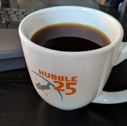 Close-in photo of a white mug filled with coffee. The mug has a decal reading "Hubble 25," with an icon of the Hubble Space Telescope.