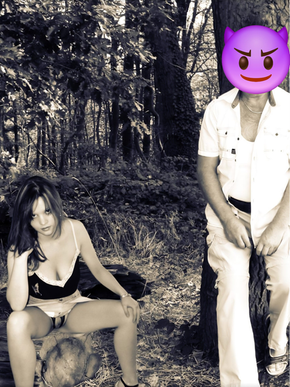Das Bild zeigt eine Frau und einen Mann in einem Wald. Die Frau sitzt auf einem Baumstamm und trägt ein schwarzes Top mit Spitzenbesatz und einen kurzen weißen Rock. Ihr Schlüppi ist ouvert und lässt einen kleinen Einblick auf ihre Charme zu. Sie hat langes, dunkles Haar und blickt nachdenklich zur Seite. Der Mann steht neben einem Baum und trägt ein weißes Hemd über einem weißen Tanktop und weiße Hosen. Sein Gesicht ist durch eine lila Teufelsgesichtsemoji verdeckt. Der Wald im Hintergrund ist dicht mit Bäumen und Laub bedeckt, und es scheint ein sonniger Tag zu sein. Das Bild ist in Schwarz-Weiß gehalten, mit Ausnahme des Emoji, das in Farbe dargestellt ist.
