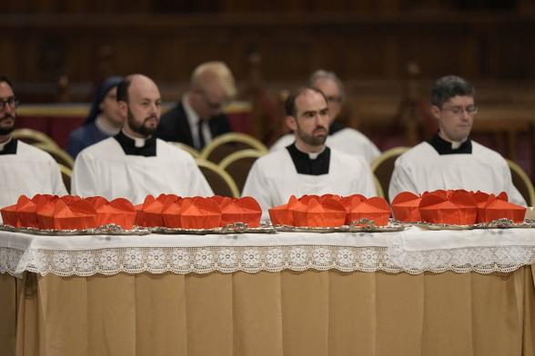 Several priests are sitting behind a table which contains trays with red caps for cardinals(?). The table is covered in a white table cloth with a lace edging.