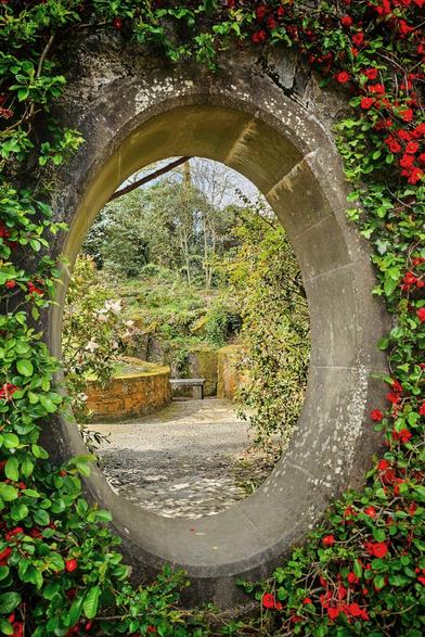 A stone oval archway is surrounded by lush green vines and vibrant red flowers, framing a view of a serene garden path with a stone bench and various plants and trees in the background.
