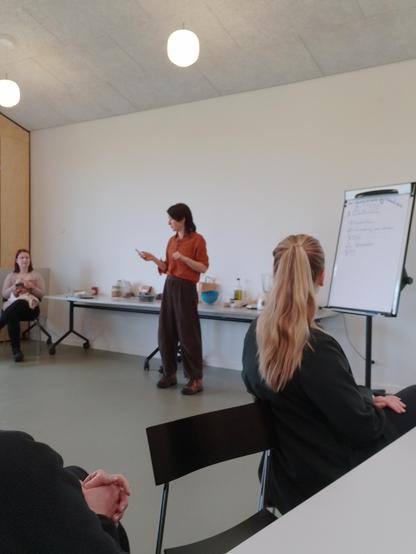 A dark-haired person in black corduroy pants and an orange shirt stands in a large room talking. Many jars and a board with agenda behind them.
People sit in the foreground. One person sits and knits and watches in the background
