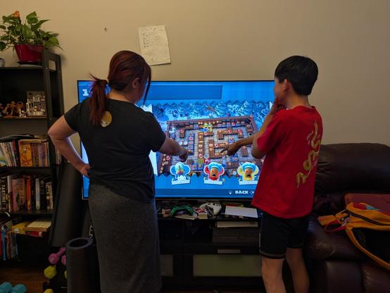 Two people (mother and son) standing in front of TV in living room, looking and pointing at the TV. Overcooked video game is being played on the TV.