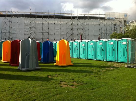 Exterior colour photo taken on a large square in Amsterdam before a local festival took place. on the right stands a row of uniformly green WC cabins lined up next to each other. On the left is a collection of free standing portable urinals in a variety of colours.