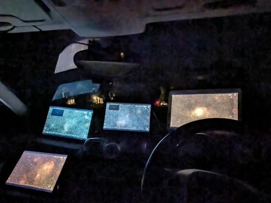 The dashboard and steering wheel of a small car, looking forward out through the windscreen. Very little can be seen outside, as there are four large tablets propped up against the windscreen.