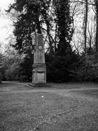 Black and white out-of-focus photo of a stone monument nestled among tall trees and dense foliage. The monument stands at the edge of a field of grass.