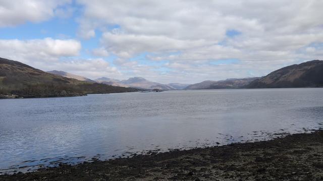 A loch with a rocky beach in the foreground and old mountains on the opposite side going back into the distance.