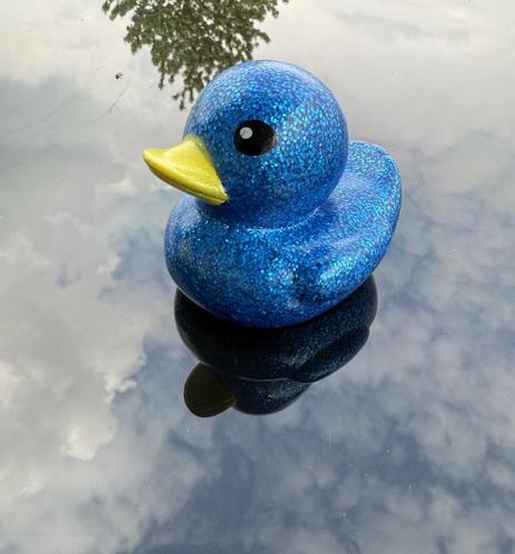 An electric blue glitter rubber duck sits atop a black glass table with blue skies and clouds reflecting around it. The duck also has a reflection.