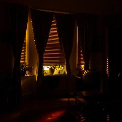 Dark photo of a living room. Yellow street light comes into the room through half-closed wooden blinds. Curtains are tied back. The light illuminates pot plants on the window ledges. The shadows light stripes across a cushion in a chair. A pool of light picks out a red carpet on the floor.