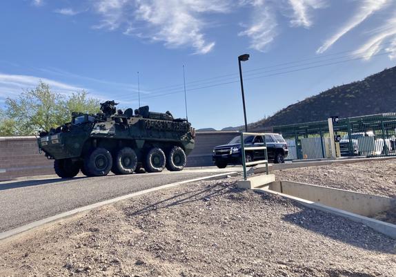 A large armored personnel carrier with eight tires prepares to leave a large Border Patrol station surrounded by a wall and fence.