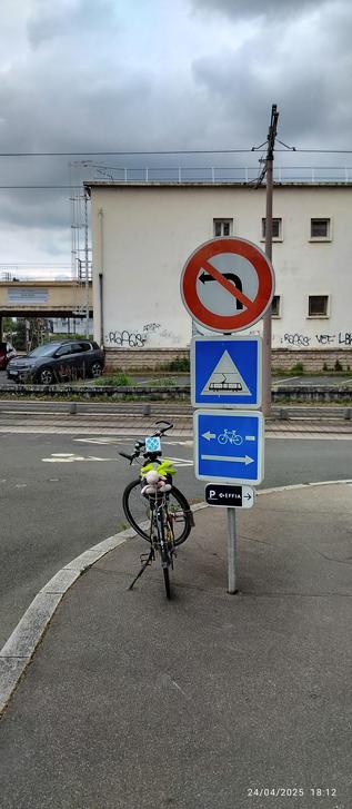 Un vélo stationne devant un panneau rond indiquant une interdiction de tourner à gauche (manque le sauf-vélo), un panneau carré indiquant le passage du tramway, un second panneau carré indiquant les sens de circulation les autos et des vélos.