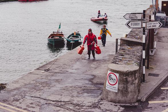 A harbor scene in Baltimore, Co Cork, Ireland showing people in waterproof gear carrying red fuel containers on a dock with small boats moored nearby, directional signs for "TOILETS", "FERRY", and "CAR PARK" visible, and a 6km speed limit sign in the foreground.