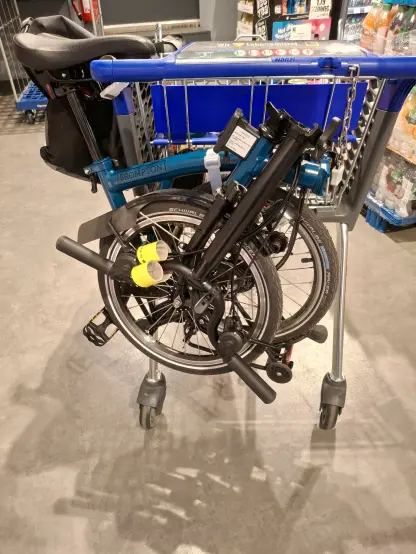 Einkaufswagen in einem Supermarkt. Am Handgriff hängt ein gefaltetes Brompton Faltrad. An der Seite hängt noch eine große Fahrradtasche.