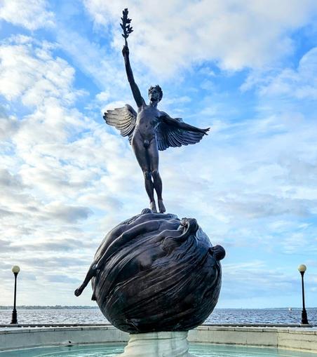 With the Saint Johns River and the big blue Florida sky as a backdrop, a view of a large bronze statue situated in a fountain, as part of a city park on the shoreline of a vast river. The statue features a winged man raising an olive branch, standing atop the globe, covered with depictions of the world's troubles.