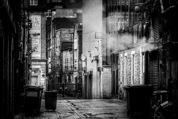 A black and white photograph shows a narrow urban alleyway with wet pavement, flanked by brick buildings and various industrial elements like pipes and bins. Light filters in from the end of the alley, creating areas of shadow and highlights.