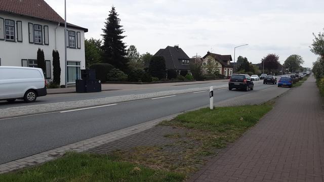 Zweispurige Straße mit MIV. Im Bildhintergrund links steht ein weiße Haus am Straßenrand. Die Straße ist gesäumt von einem rot gepflasterten Fußweg mit Zusatzschild 1022-10, welches nicht auf dem Foto zu sehen ist (zur Erklärung der Verkehrsführung). Dazu teilweise straßenbegleitend Längsparkplätze. Auf der gegenüberliegenden Straßenseite vor dem Haus steht auf einem dieser Parkplätze ein schwarzer, fast menschengroßer Kasten. Es handelt sich um einen mobilen Blitzer, der in beide Richtungen blitzt. Die Parkplätze davor und dahinter sind alle frei, der Blitzer ist wegen der geraden Straße weit im Voraus zu erkennen.