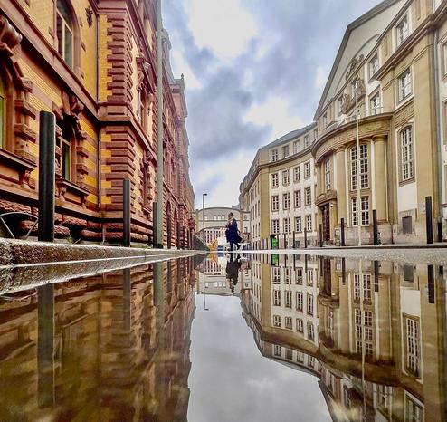 Das Bild zeigt eine beeindruckende Stadtansicht mit historischen Gebäuden, aufgenommen aus einer sehr niedrigen Perspektive – knapp über einer großen Pfütze auf der Straße. Die Szenerie wirkt besonders eindrucksvoll, weil sich die Gebäude und der Himmel fast perfekt in der Wasseroberfläche spiegeln.

Auf der linken Seite steht ein altes, rotbraunes Gebäude mit kunstvoller Fassade. Die rechte Seite zeigt ein helleres Gebäude mit runden und eleganten Formen. In der Bildmitte, weiter hinten, sieht man einen modernen Verbindungsbau, der zwei Gebäudeteile über der Straße verbindet.

Der Himmel ist wolkenverhangen und wirkt grau-blau. Zwei Menschen stehen in der Mitte des Bildes auf dem nassen Pflaster – eine Person scheint zu fotografieren oder auf ihr Smartphone zu schauen. Die Farben sind trotz des trüben Wetters kräftig, und die Perspektive sowie die Spiegelung erzeugen eine fast surreale, kunstvolle Stimmung.