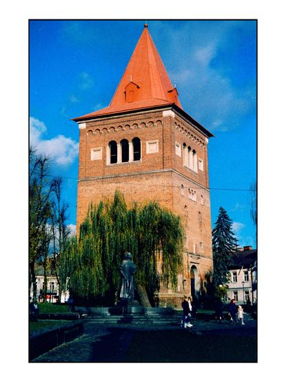 The image features a tall brick tower with a distinctive red conical roof, set against a clear blue sky with a few scattered clouds. The tower has a square base with arched windows on the upper level, and a large arched entrance at the ground level. In front of the tower, there is a statue of a person on a pedestal, surrounded by a weeping willow tree. The area around the tower is a park-like setting with people walking and sitting, and other buildings visible in the background. The overall scene is well-lit, suggesting it is a sunny day.