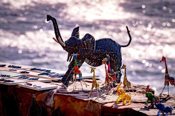 The image features wire and bead crafts shaped like various animals, including an elephant and several giraffes, displayed on a surface near water. The crafts are colorful and detailed, with the water in the background sparkling in the light.