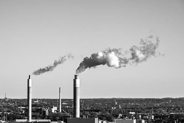 Black and white photograph of an industrial landscape, featuring several tall smokestacks rising above a sprawling city. Dense plumes of smoke drift from the chimneys, highlighting the contrast between heavy industry and the surrounding urban environment.