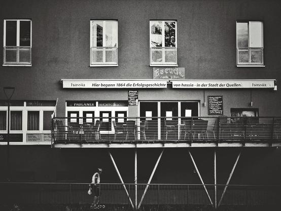 The landscape-orientated black and white image shows the facade of a building. The upper part of the building has four rectangular windows with white frames and a silver-colored structure beneath each. Below these windows is a white awning with black lettering. A balcony or terrace area extends from the building beneath the awning, featuring metal railings and outdoor seating with tables and chairs. Behind the balcony furniture are large glass doors and windows. To the left of the balcony, there are additional windows. A lamppost is present on the far left side of the image. The lower part of the building's facade is visible beneath the balcony, with several small rectangular windows. In the foreground, there is a metal fence or railing and a strip of green grass. A person wearing a colorful top, black shorts, and a helmet is on rollerblades in the foreground, moving horizontally. Text is visible on the building, including "hassia" and "PAULANER".