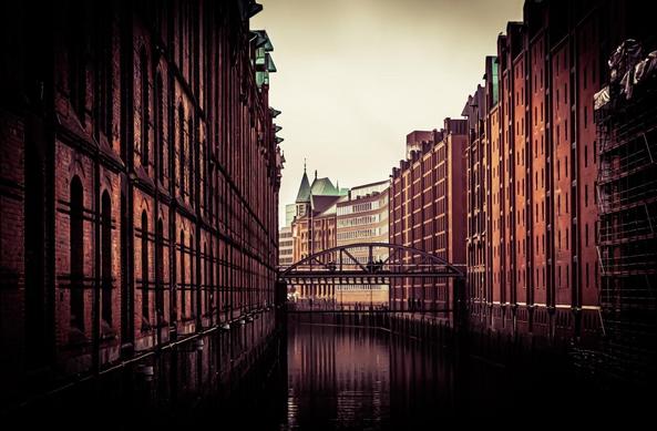 The image depicts a canal flanked by tall, brick-clad buildings. There is a bridge spanning across the canal in the mid-ground. The architectural style and setting suggest an urban area with historical buildings, possibly in a city known for its waterways. The atmosphere is moody with a somewhat vintage or sepia-toned effect.