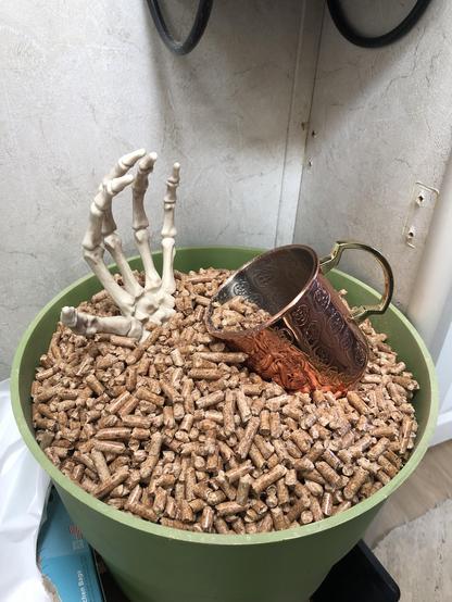 A large green planter is filled with wood pellets (for the toilet). A skeletal hand rises from them, as if from a grave. It overshadows a copper mug being used as a scoop, as if to grab it.