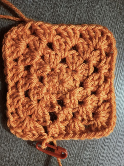 Labor de ganchillo de lana naranja sobre mesa de madera gris.
Tiene forma cuadrada con las esquinas redondeadas y se le abren una serie de huecos de manera que recuerda a un tablero de ajedrez, en el que los huecos serían las blancas y los grupos de puntos las negras.

Parte de un anillo mágico en torno al que he tejido 12 puntos altos.
En la siguiente fila, he metido 3 puntos altos, una cadeneta y 3 puntos altos en un mismo hueco, creando una esquina redondeada. El proceso se repite 3 veces más, en huecos a 3 espacios de distancia, creando así las 4 esquinas.
En la tercera fila, en el hueco de la cadeneta de las esquinas se le meten otros 3-1-3 puntos, y en los huecos de la segunda fila que no son esquina se le meten 3 puntos altos.
La cuarta fila sigue la misma lógica: grupos de 3 puntos en los huecos que no son esquina y 3-1-3 en los que sí.

El último punto está asegurado con un diminuto imperdible rojo.