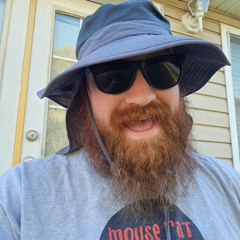 Selfie of a very thick reddish bearded white man wearing sunglasses and a floppy grey hat with a neck flap and a grey t-shirt that reads "Mouse Rat" in red text within a portion of a black.