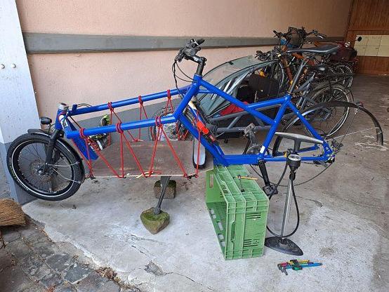 Das Bild zeigt ein blaues Lastenrad mit roten Spannbändern, das auf einem selbstgebauten Montageständer steht. Der Ständer besteht aus einer grünen Kunststoffkiste unter dem Rahmen und zwei Steinen unter dem Doppelständer, die das Fahrrad anheben und stabilisieren. Am Lastenrad ist das Hinterrad ausgebaut. Im Hintergrund sind weitere Fahrräder zu sehen. Auf dem Boden befinden sich einige Werkzeuge und eine Standluftpumpe.