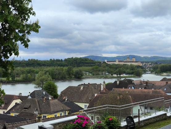 Blick auf viele dunkelrote Ziegeldächer, dahinter ein Fluss und auf der anderen Flussseite ein großes barockes Stiftsgebäude.
