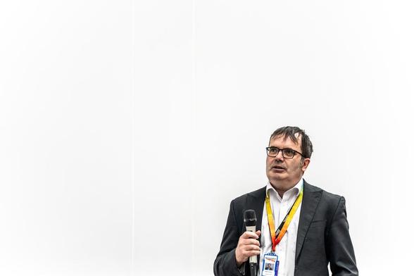 A picture of a bloke in a suit, standing in front of a white background, holding a microphone with a rainbow lanyard and staring into the middle distance