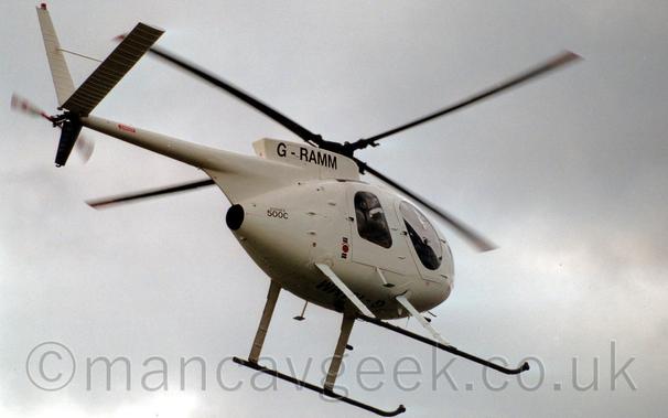 Low side view of a white helicopter flying from left to right at a very low altitude.
The helicopter is all white, with the registration "G-RAMM" on the side of the engine cowling, repeated under the belly between the landing skids.
There are smaller, red, "Hughes 500C" text on the side of the rear fuselage, under the tail boom.
A pair of doors on the side of the fuselage have large windows, with people vaguely visible inside.
Grey sky fills the background.