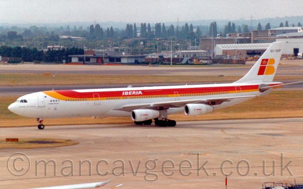 Side view of a white, 4 engined jet airliner taxiing from right to left.
There is a yellow, orange, and red strip coming down from over the top of the forward fuselage and running backwards along the body, with white "Iberia" titles over the wings.
There are a pair of large letters on the tail - a red, lower-case "I" with a gold crown on the dot, and a yellow upper-case "B", with the registration "EC-GUP" at the top of the tail in black.
The white engine pods under the wings have small, red, "Iberia" titles in the middle.
Grey concrete apron fills the foreground, with yellowing grass and a runway lead up to more apron and buildings in the distance, with trees beyond, under grey sky.