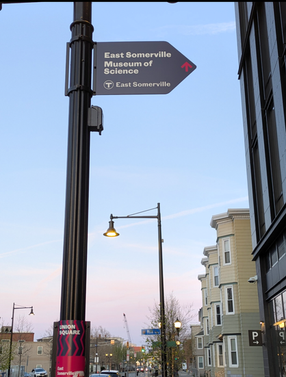 Directional sign on a light post with the text "East Somerville Museum of Science" (those are two separate things but there's no indication of that on the sign), with smaller text reading "T (MBTA) East Somerville" closer to the bottom of the sign. To the right of the larger text there's a small red arrow (poor contrast with the dark blue background of the sign). The arrow points up to indicate these attractions are both straight ahead, but in reality only one of them is straight ahead, the other would be to the viewer's left. And to make it even worse, the sign itself *is in the shape of an arrow that points to the right*!!