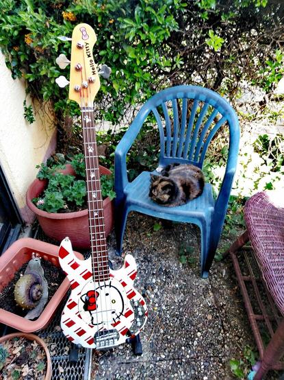 A white and red Hello Kitty themed bass guitar. Tuned BEAD, the bass itself is a modified Harley Benton passive Stingray clone. The bridge, pickup, electronics, and tuners have all been replaced, down to the knobs and jacks. The wide neck is still original, and perfectly serviceable.
The bass is sitting outside on a terrace, next to a blue plastic chair containing a sleeping tortoiseshell cat (RIP, Cleo), and a flower pot containing a giant spiked snail. Shrubbery is in the background.