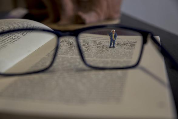 bild im querfromat, in der bildmitte eine brille, die auf einem buch liegt.
durch das brillenglas wird in dem bereich die schrift scharf. im rechten brillenglas steht eine kleines männchen mit brille, anzug und krawatte und hält sich den rücken
