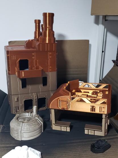 3d printed rectangular structures in copper and bronze filament. One of a 3 story tall apartment building with a 3rd floor balcony, the other is an open walled bar.