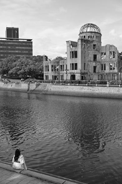 A black and white photograph of the A-bomb dome from across the river. A young woman in a white dress sits by the riverside looking at it.