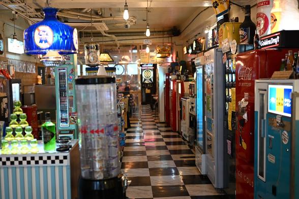 A view into the retro gaming bar museum. Colorful qntique drink and food dispensers line the checkered floor corridor. The aesthetic is quite 1950s-80s.