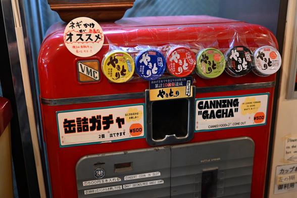 A closeup of a bright red food dispenser. It has a bunch of cans attached to it with labels in Japanese, and it says "Canning Gacha. I'm looking forward to seeing what's in it. Canned food x 2 come out" 
The types are a mystery to me.